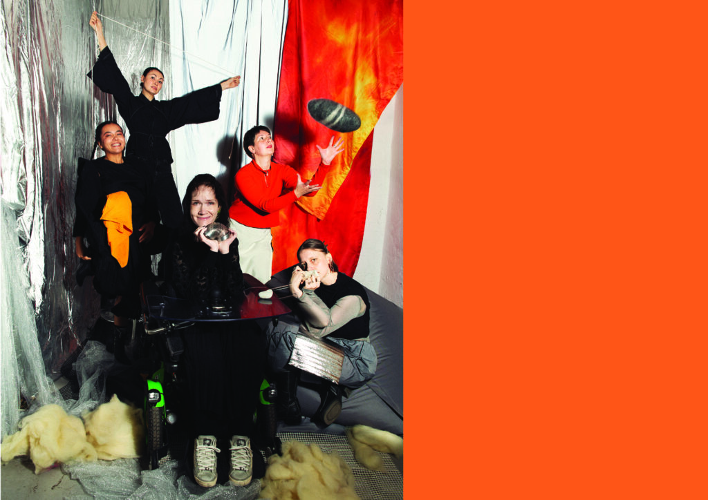 Image description: The photo shows the five performers of Dissolution: Miriam Cochanski, Kira Shmyr, Jeanna Kolesova, Ziliä Qansurá and Victoria Sarangova. In the foreground, in the center, is Miriam. She is a white woman sitting in her wheelchair. Miriam has long, brown, curly hair, wears a black blouse, a short black skirt, black leggings, and grey shoes. She is smiling directly at the camera while holding a silver slinky. On the right is Kira, sitting on a grey beanbag. Kira is a white non-binary person with light brown hair styled in a bob. They are wearing grey trousers, a grey mesh long-sleeved t-shirt, a black sleeveless t-shirt, and black boots. Kira holds a grey stone in their hands, covering part of their face, with only their eyes visible. Jeanna stands behind Kira. They are a white non-binary person with short brown hair, wearing black trousers, an orange jumper, and a beige apron. Jeanna is throwing up a soft, grey felt wool stone, looking at it with concentration as it flies over Kira's head. Behind Miriam, on the left, is Ziliä, a BIPOC woman with long black hair braided into two pigtails. She is wearing a black dress and black boots, with an orange scarf hanging from her waist. She is jumping and laughing. Victoria is standing on a ladder behind everyone. She is a mixed Asian (Oirat)/Armenian woman with black hair in a ponytail, wearing black trousers and a black blouse with wide sleeves. She is stretching a thin white rope in opposite directions with a thoughtful expression on Victoria's face.