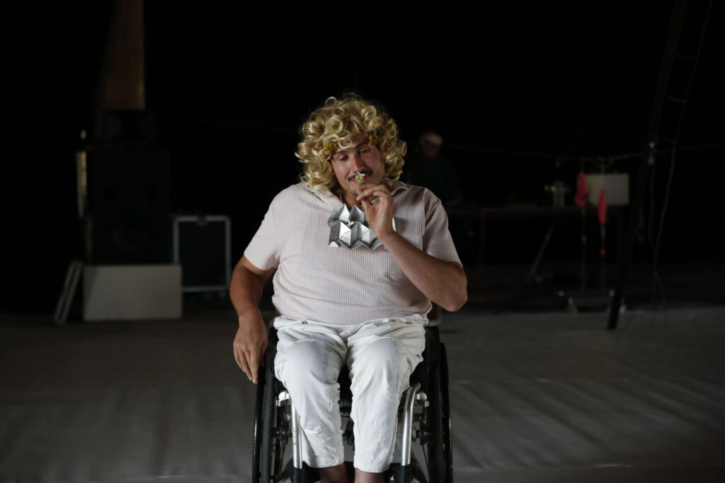 Performer*in mit blonder Lockenperücke sitzt im Rollstuhl auf dunkler Bühne und hält eine kleine Blume an die Nase.